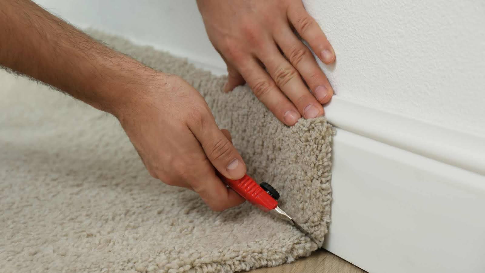 Hands using a utility knife to trim carpet along a wall edge